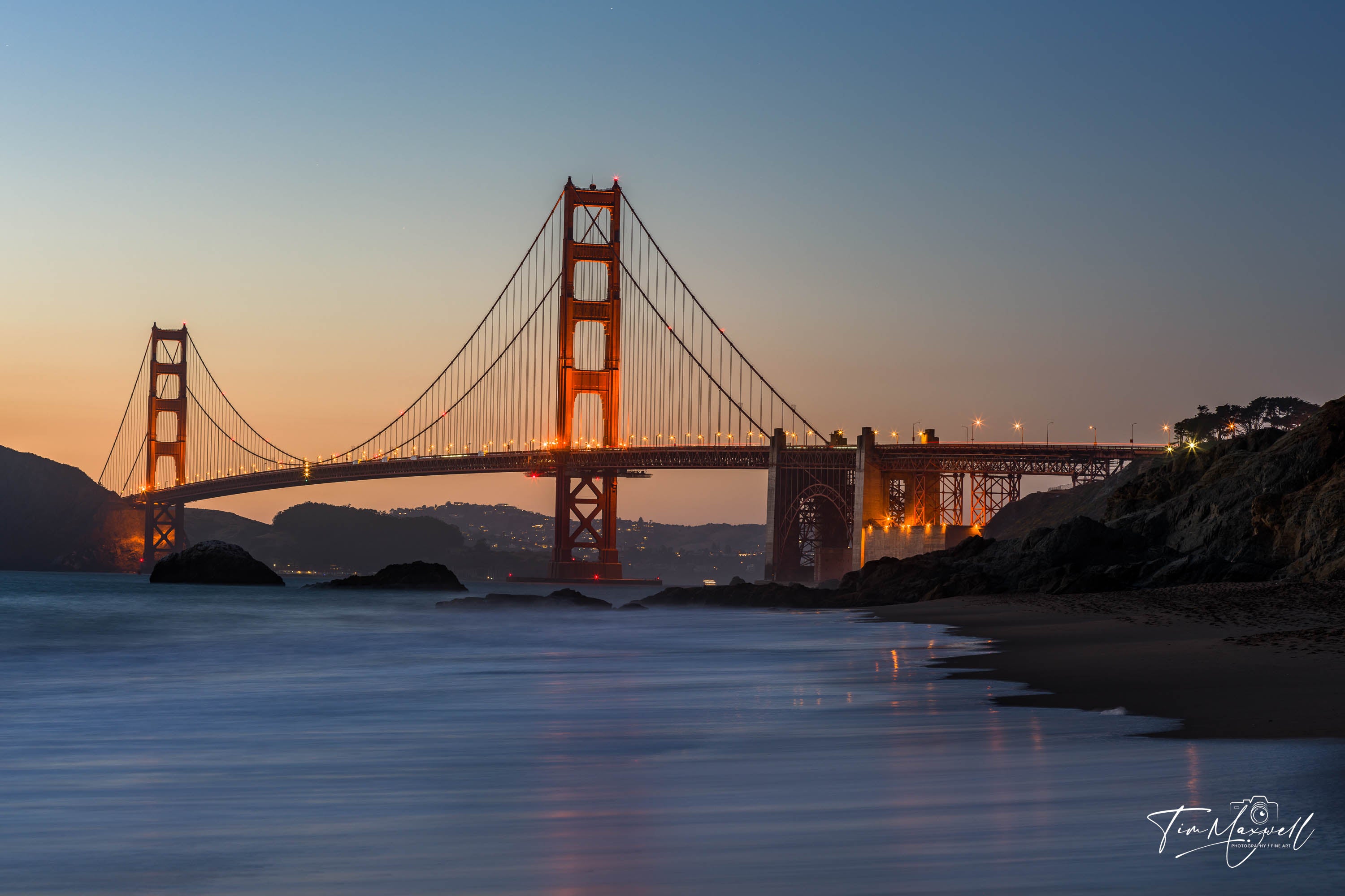 Golden Gate Bridge illuminated at night, viewed from Baker Beach with soft waves and a peaceful shoreline