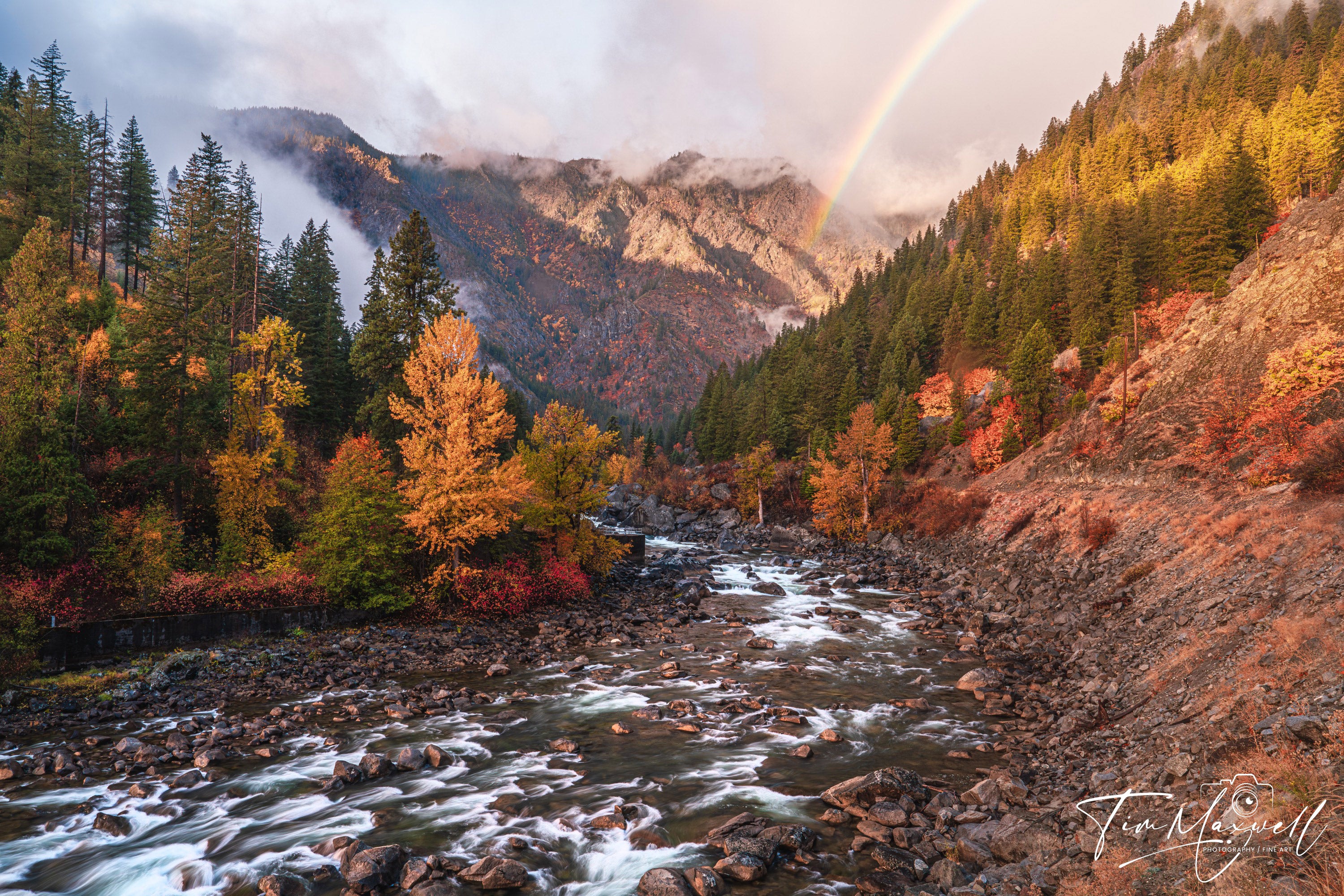 Autumn foliage along Tumwater Canyon with a vivid rainbow arching over misty mountains, fine art print by Tim Maxwell.