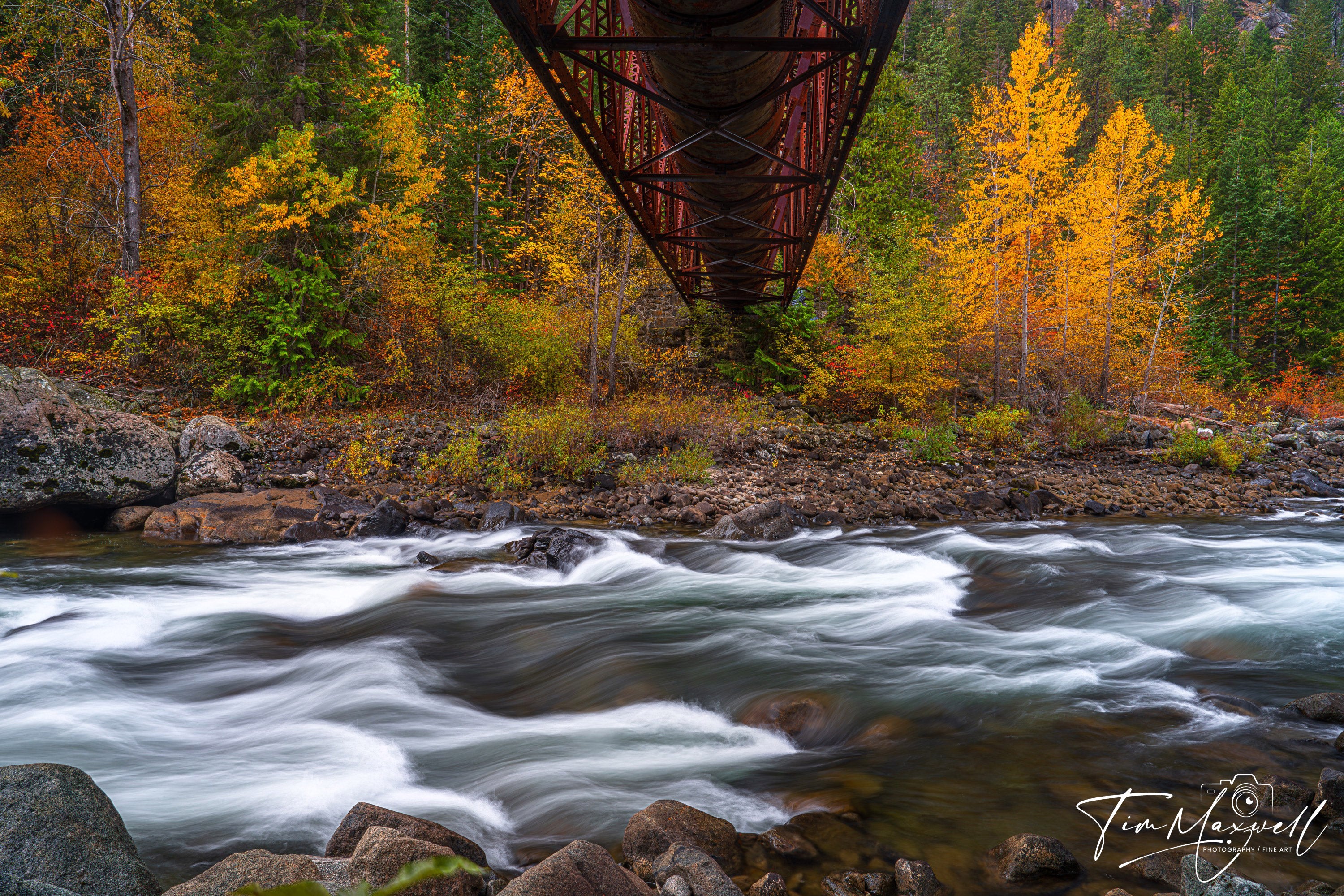 Long-exposure of the Wenatchee River flowing under a rustic iron bridge, surrounded by peak autumn foliage in Tumwater Canyon.